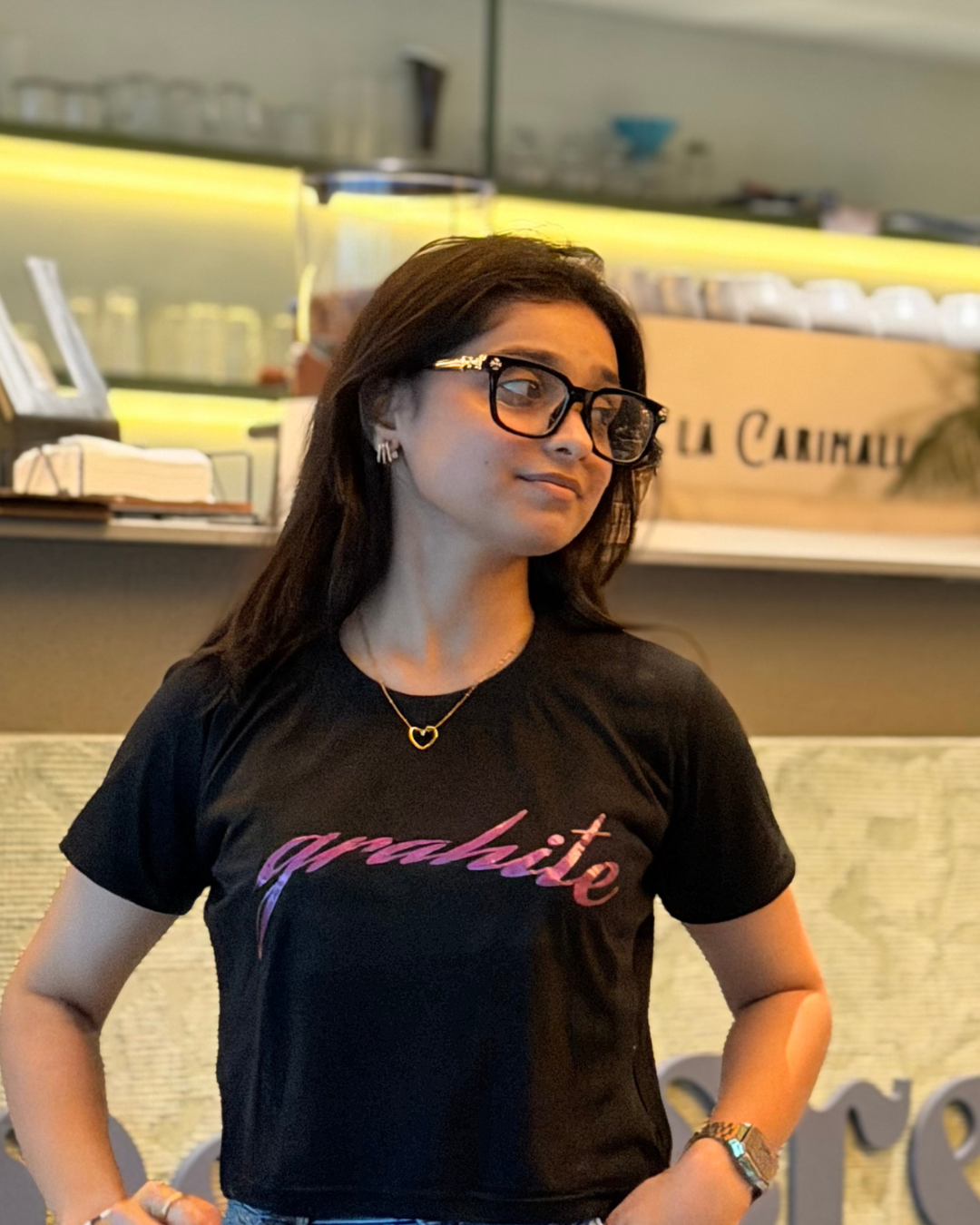 A woman wearing a black t-shirt with 'grahite' printed in pink script across the chest, standing in a cafe environment.