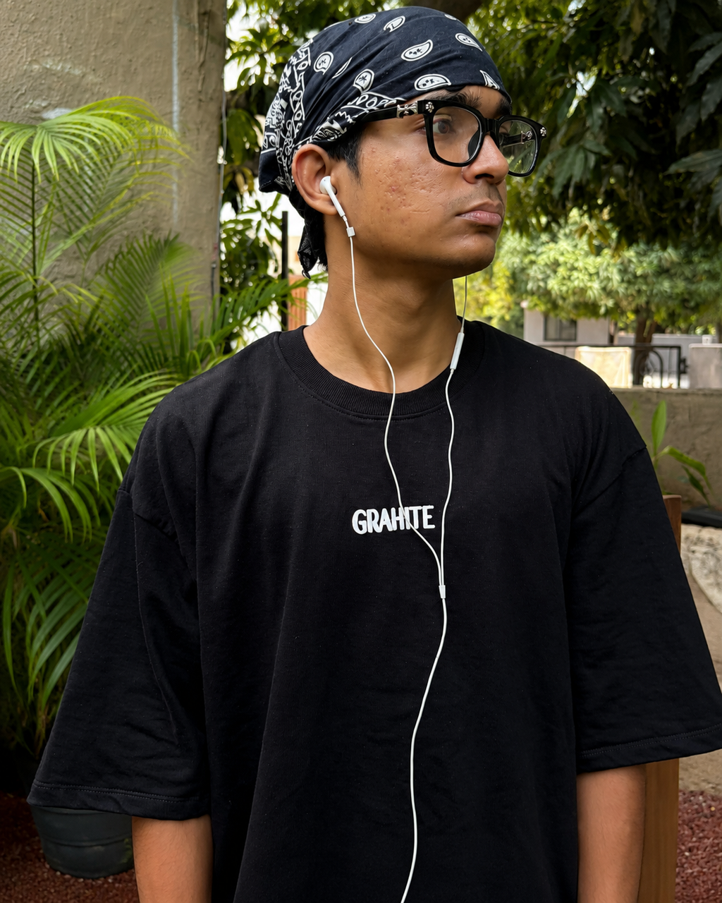 A man wearing a black short-sleeve t-shirt with 'GRAHITE' printed in white on the chest, a black and white paisley bandana, and glasses, standing outdoors with plants in the background.
