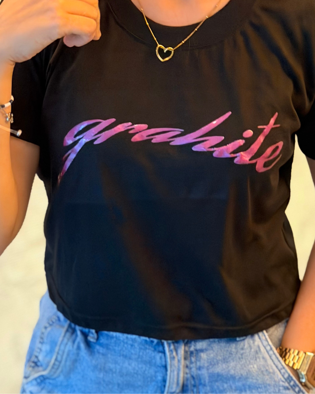 A person wearing a black short-sleeve CROP TOP with the word 'grahite' printed across the chest in a pink-to-purple gradient script, paired with light blue jeans and a gold heart necklace.