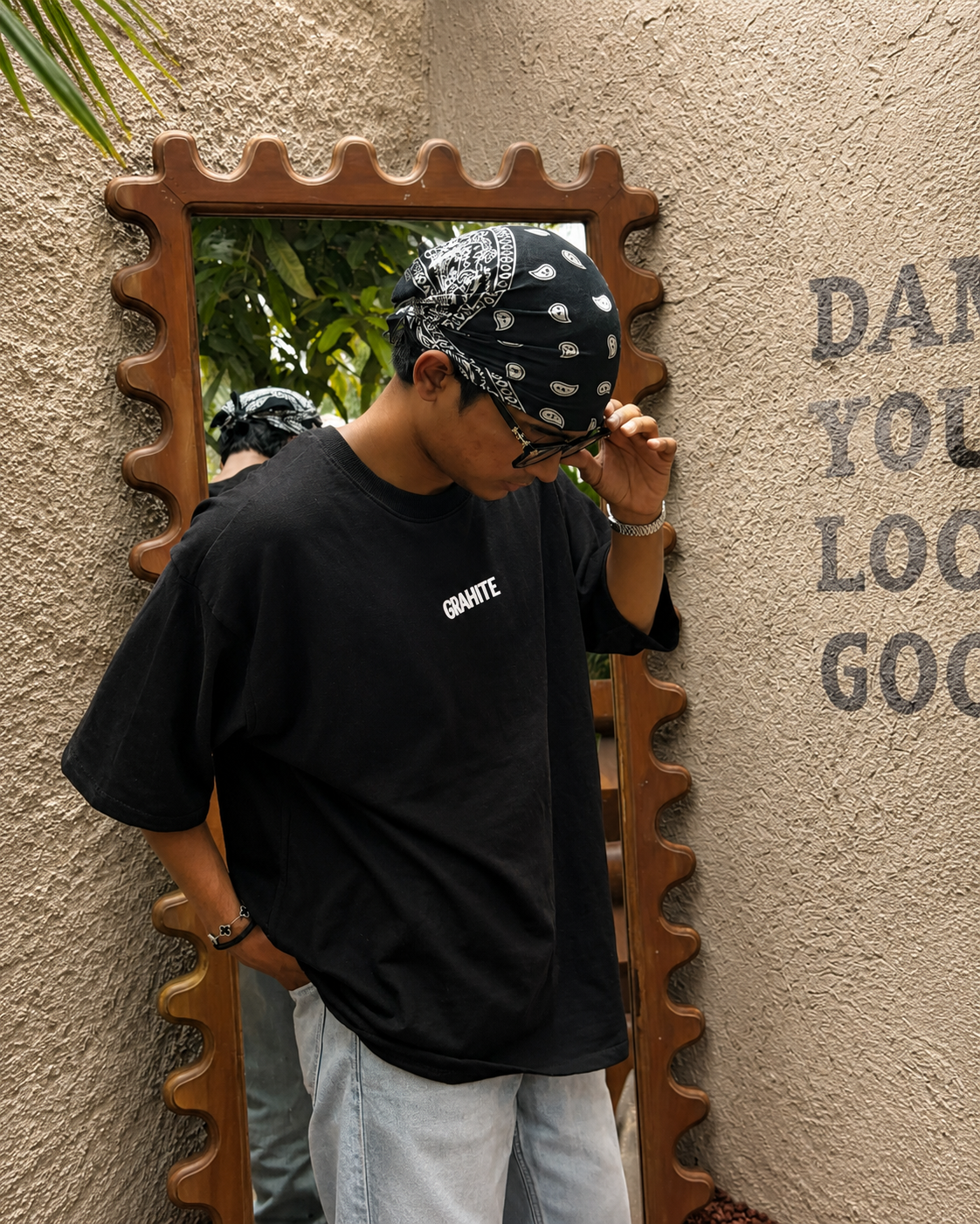 A person wearing a black oversized t-shirt with a white 'GRANITE' logo on the chest, paired with a black and white paisley bandana and light blue jeans, posing in front of a decorative wooden mirror against a textured beige wall.