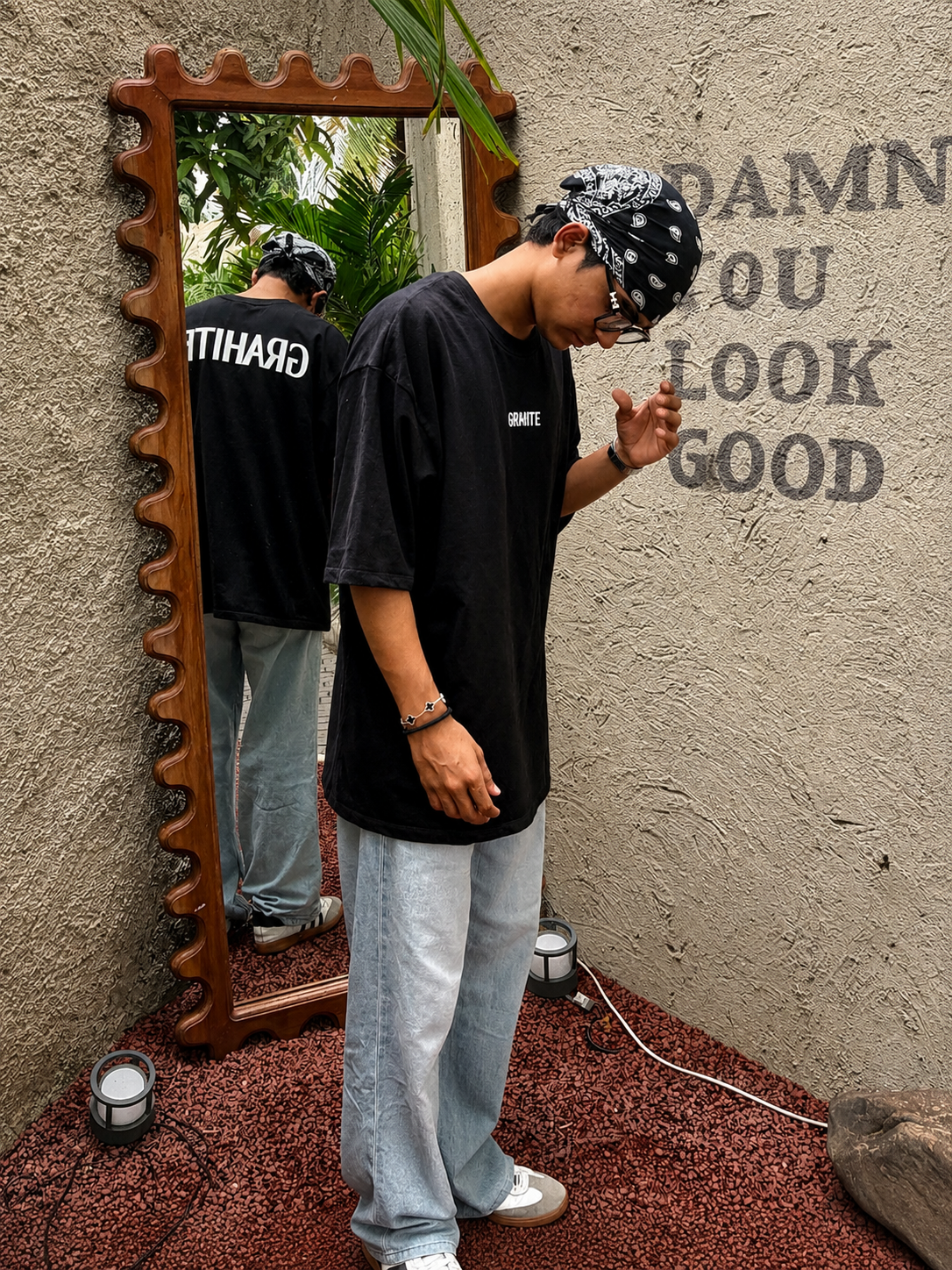 A person wearing a black oversized T-shirt with white text, light blue jeans, and a black-and-white patterned bandana stands in front of a decorative wooden mirror, looking down. The mirror reflects the back of the T-shirt, which has white text.
