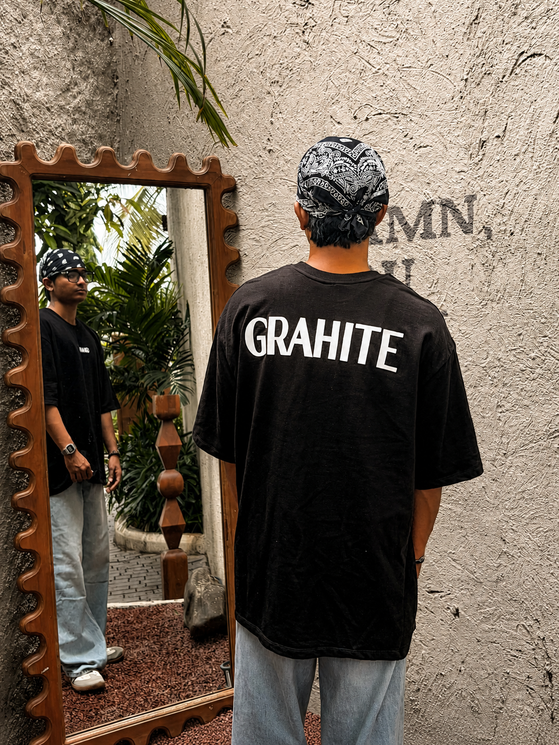 A man stands with his back to the camera wearing a black oversized t-shirt with 'GRAHITE' printed in bold white letters on the back, paired with light blue jeans and a black-and-white patterned bandana. He is positioned in front of a textured stucco wall and a decorative wooden-framed mirror that reflects his profile.