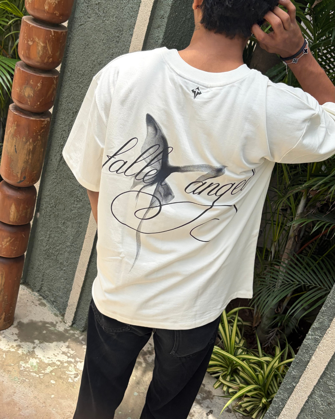 A person shown from behind wearing an oversized white t-shirt with a black graphic print of a shark and the words 'hale' and 'ango' in cursive script, paired with black jeans, standing outdoors next to a wooden post and green plants.