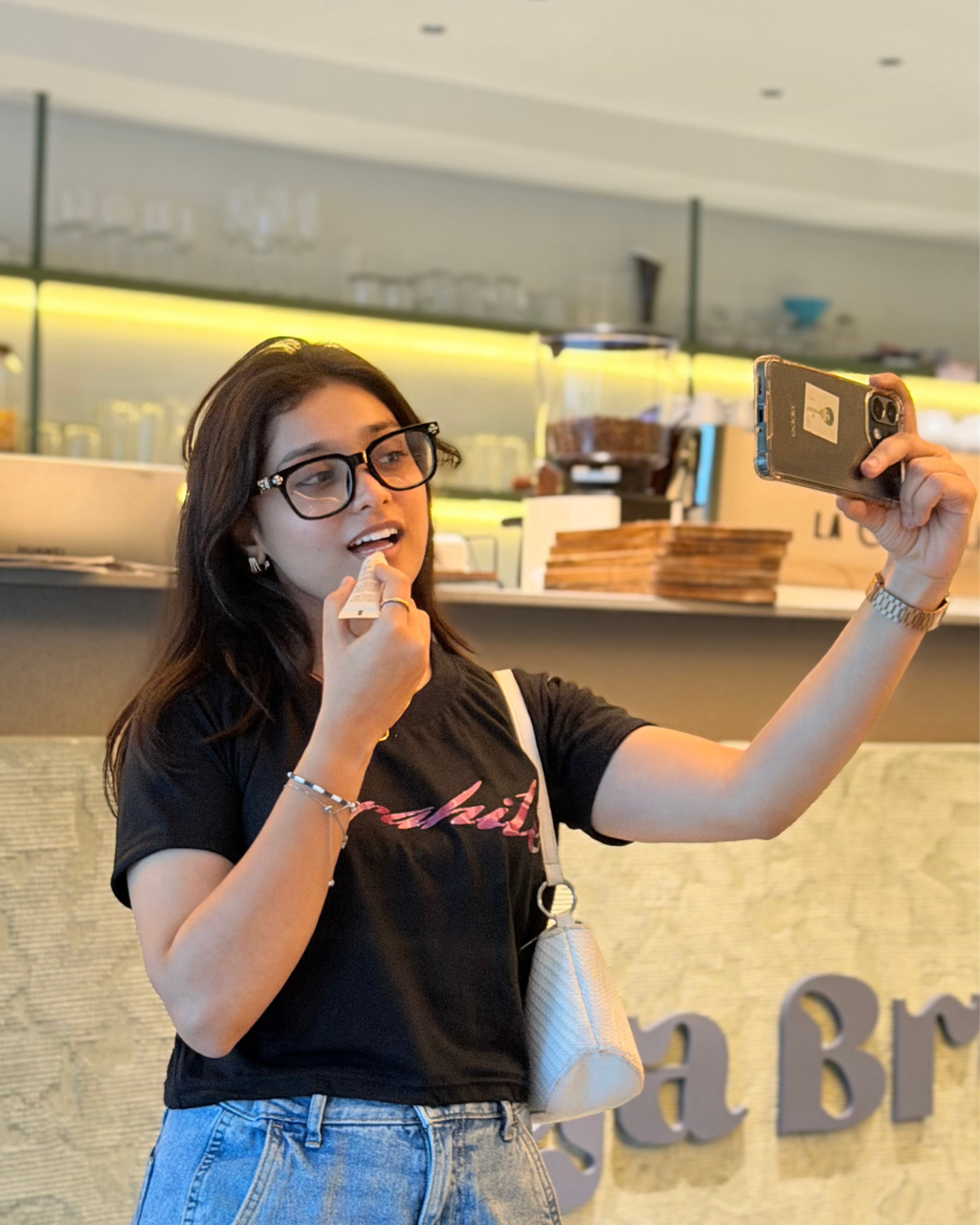 A woman wearing a black graphic t-shirt and blue jeans takes a mirror selfie in a cafe setting, holding a smartphone in one hand and applying lip product with the other.