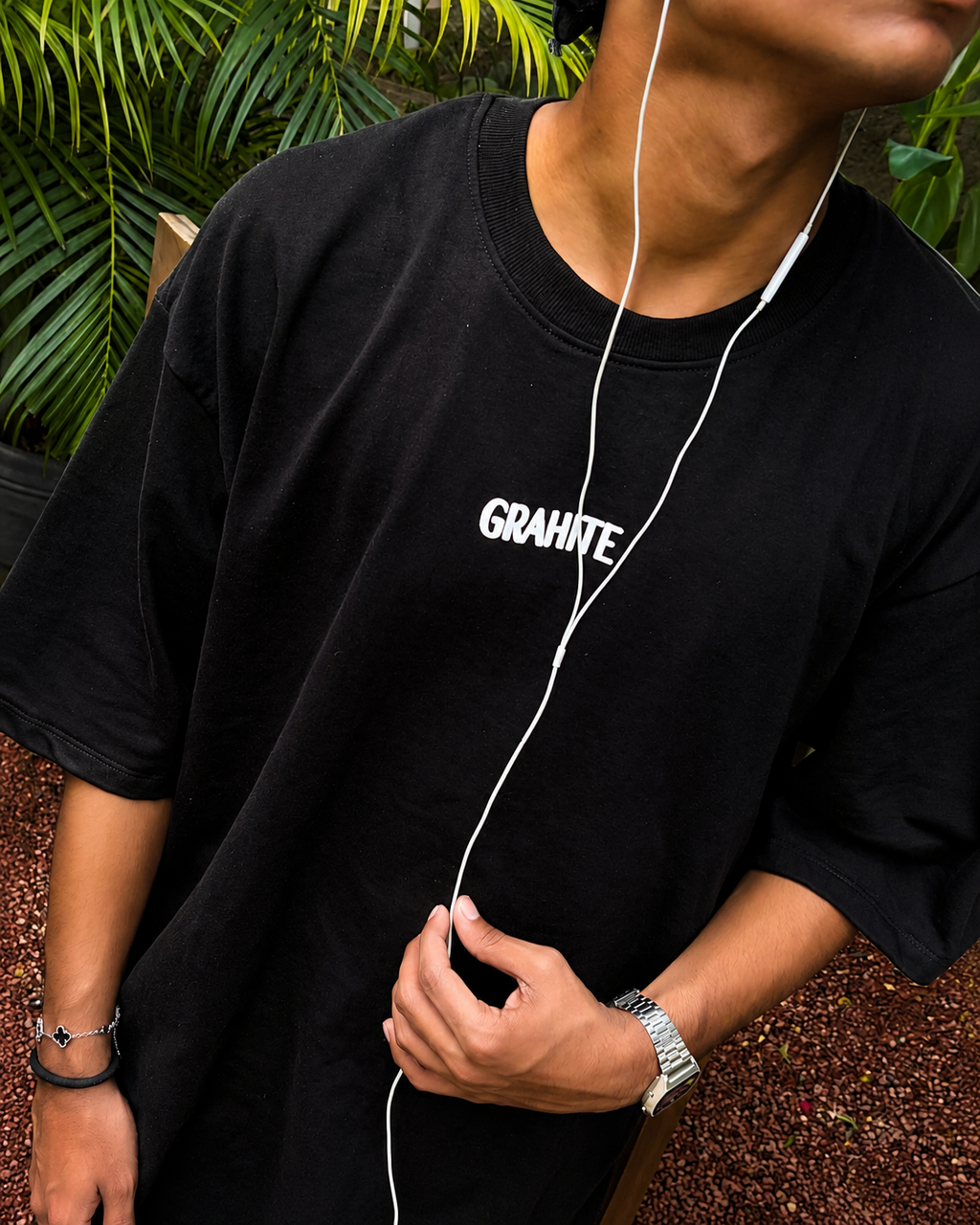 A person wearing an oversized black t-shirt with the brand name 'GRAHITE' printed in white on the chest, styled with white wired earphones.