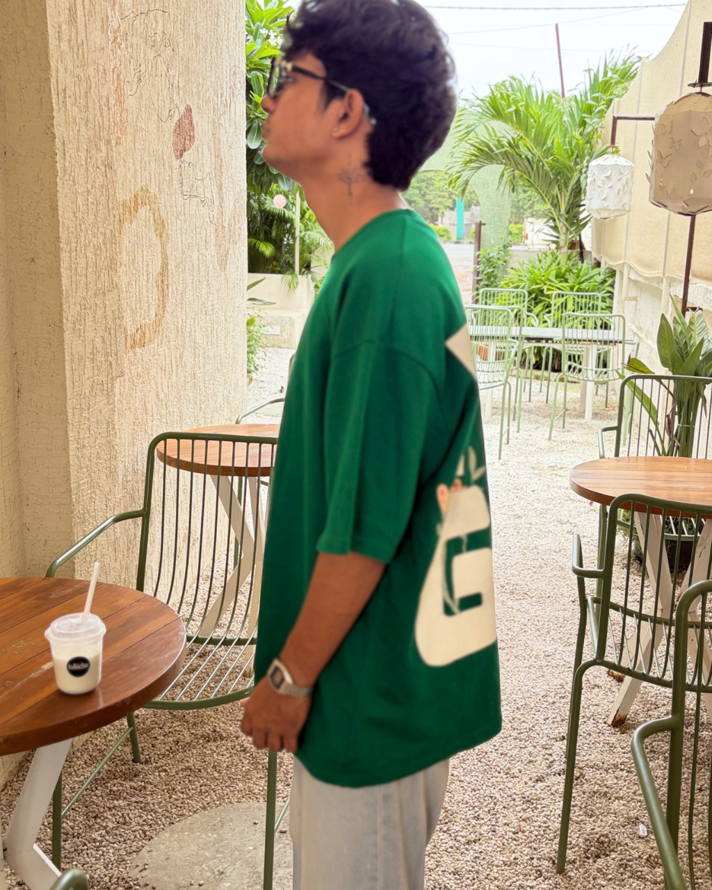 A man wearing a green oversized t-shirt with a large cream graphic on the back, standing in an outdoor patio setting with tables and chairs.