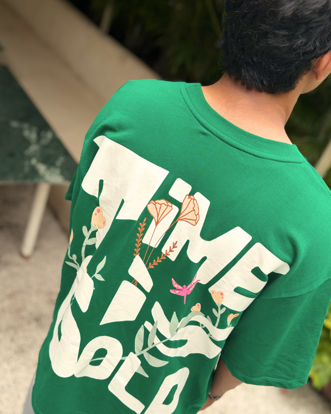 A man viewed from behind wearing a green t-shirt with a large white graphic print reading 'TIME FOR GOLD' decorated with botanical illustrations.