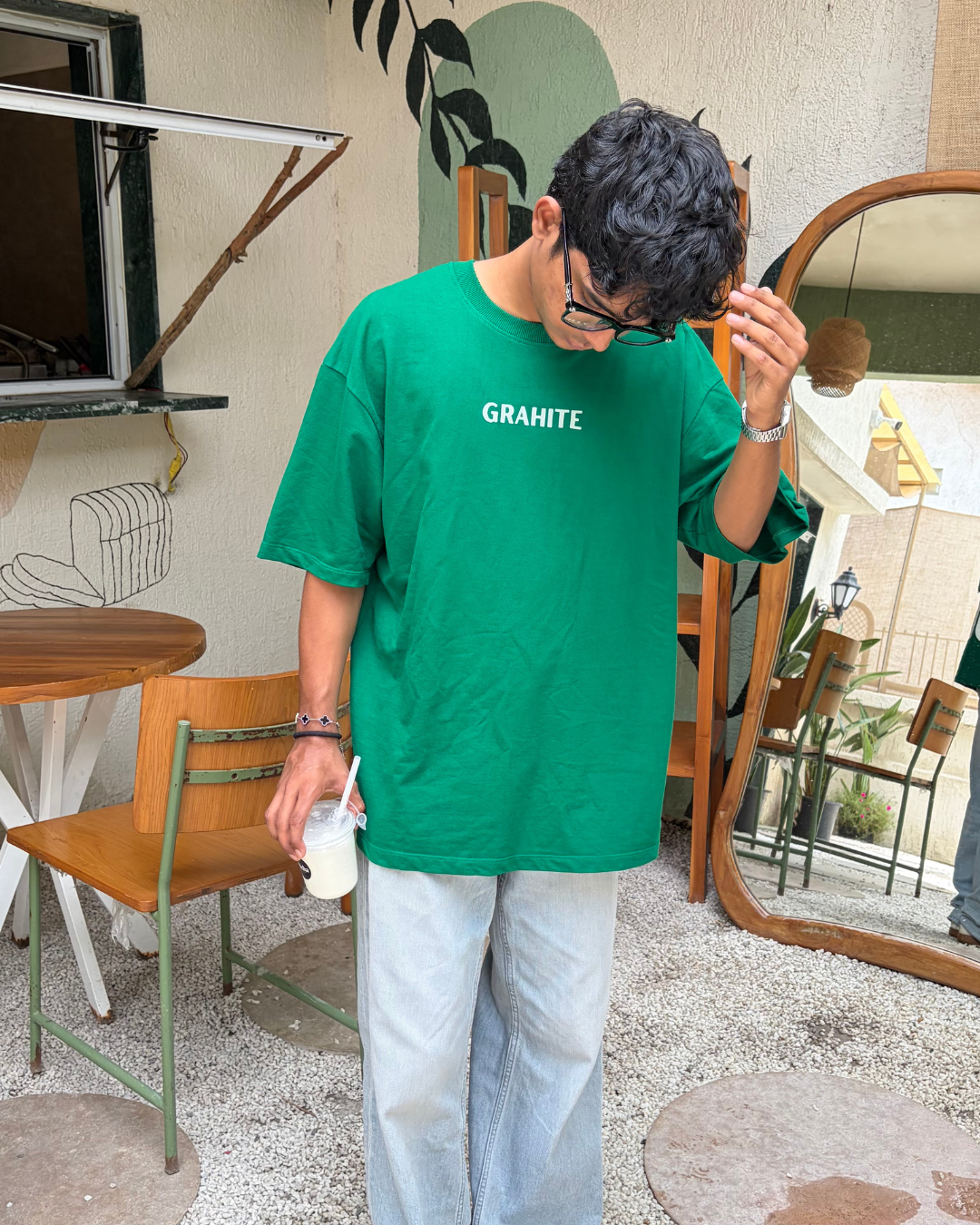 A man wearing an oversized bright green t-shirt with 'GRAHITE' printed in white across the chest, paired with light gray jeans, standing in a cafe setting.