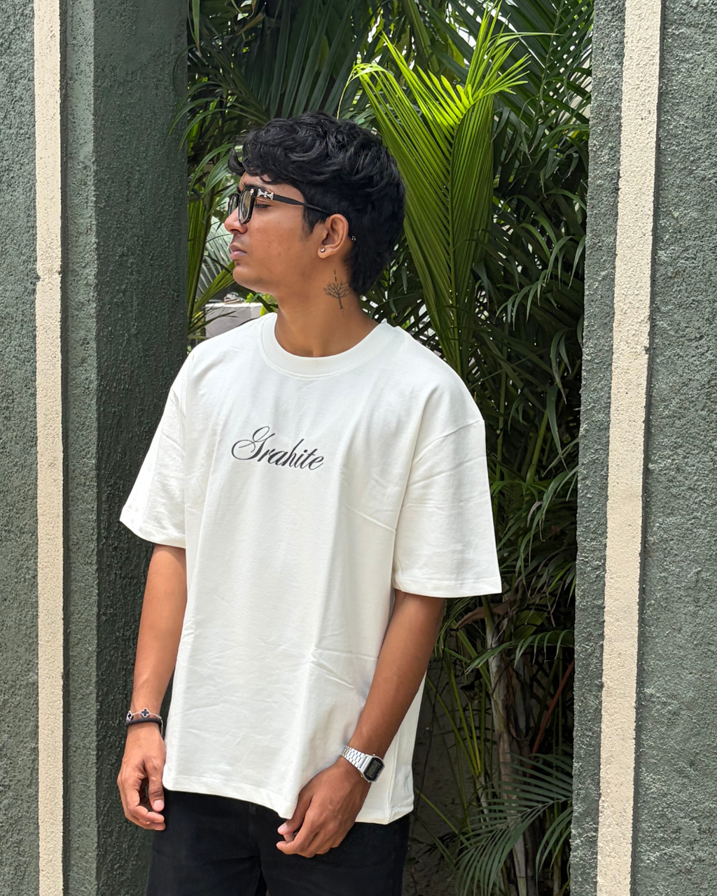 A man wearing a white oversized t-shirt with 'Grahite' printed in black cursive on the chest, standing in front of a textured green wall with palm leaves.