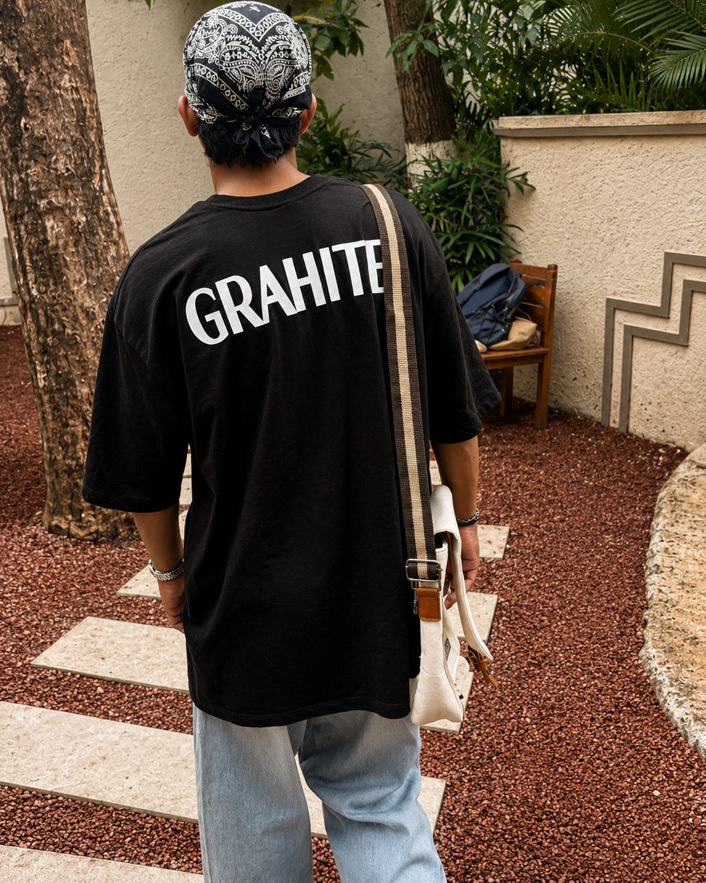 A man viewed from behind wearing a black oversized t-shirt with 'GRAHITE' printed in bold white letters on the back, paired with light blue jeans, a black and white patterned bandana, and carrying a cream-colored shoulder bag.