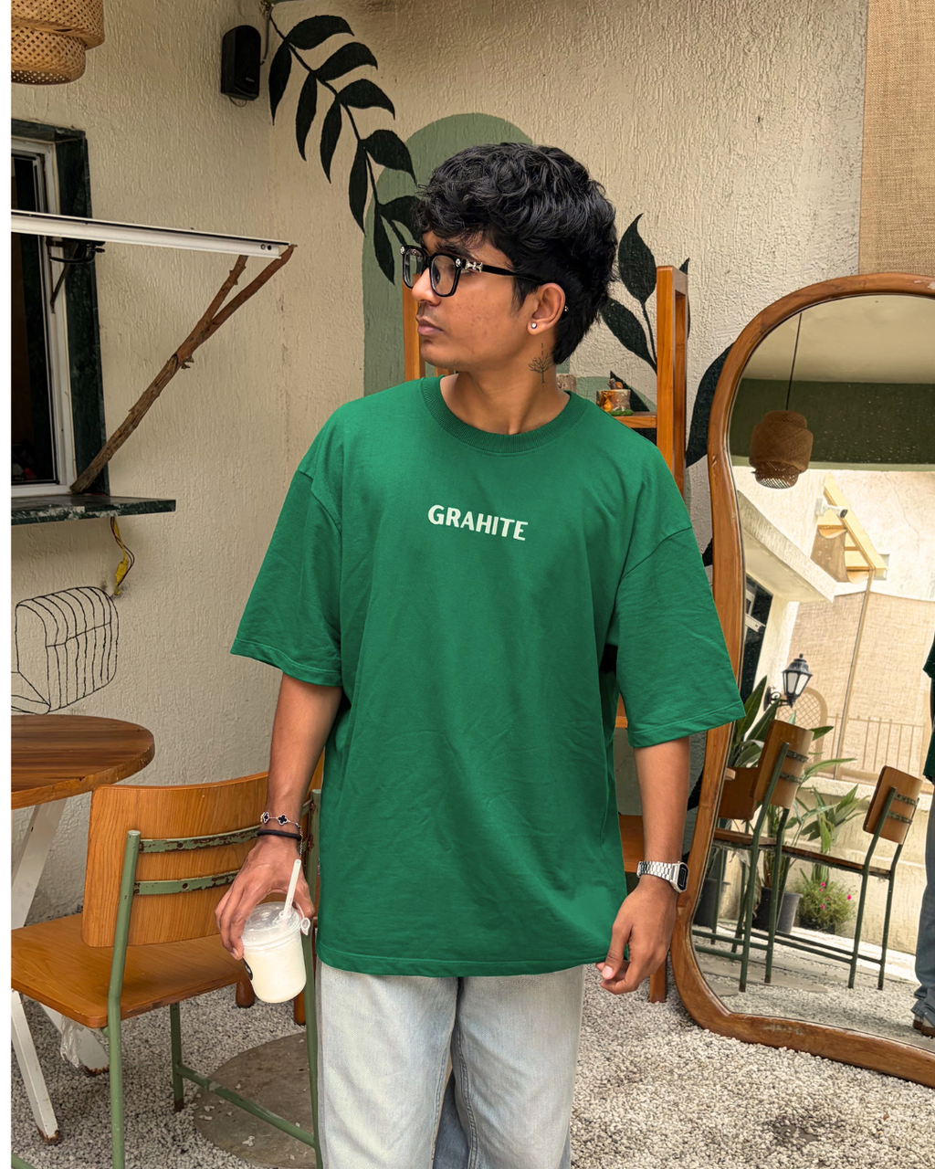 A man wearing a green oversized t-shirt with the brand name 'GRAHITE' printed in white on the chest, paired with light gray pants, standing in a cafe-like environment.