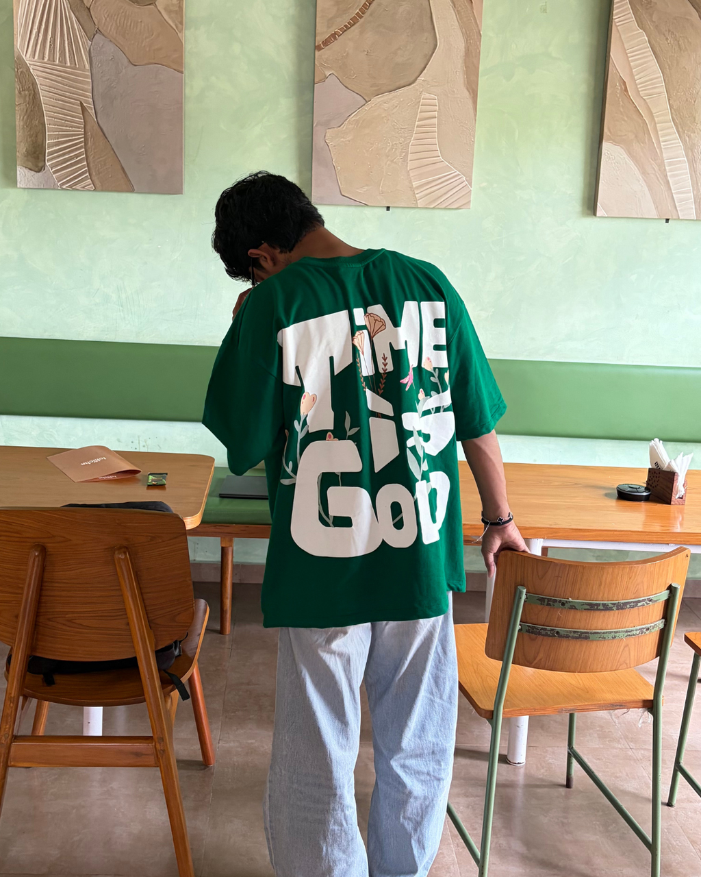 A person standing in a cafe wearing an oversized green t-shirt with a large white graphic print on the back that reads 'TIME IS GOD'.