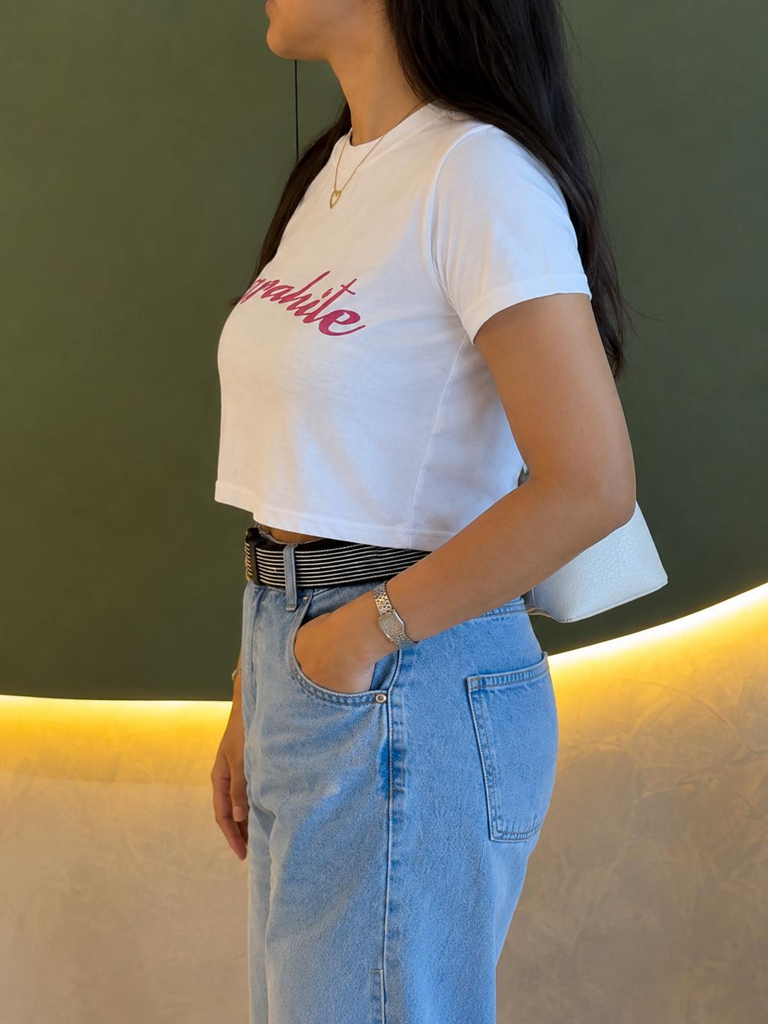 A woman wearing a white short-sleeve crop top with pink script text across the chest, paired with light blue jeans and a black-and-white striped belt, standing indoors against a dark green wall with warm lighting.