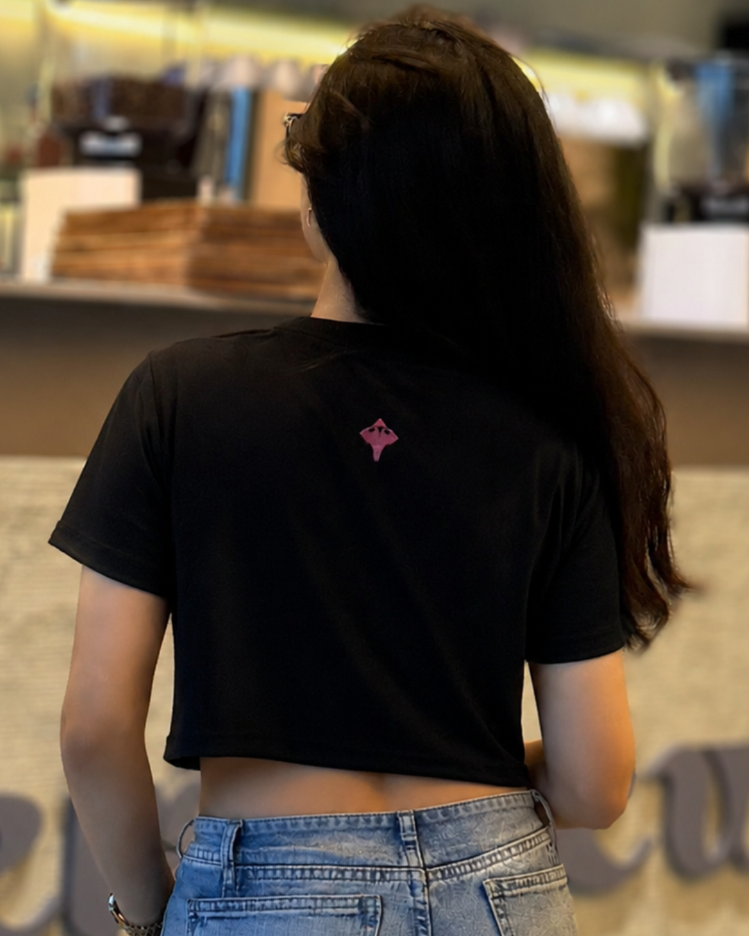 A woman viewed from behind wearing a black short-sleeve crop top with a small pink geometric logo on the upper back, paired with light blue jeans in a cafe setting.