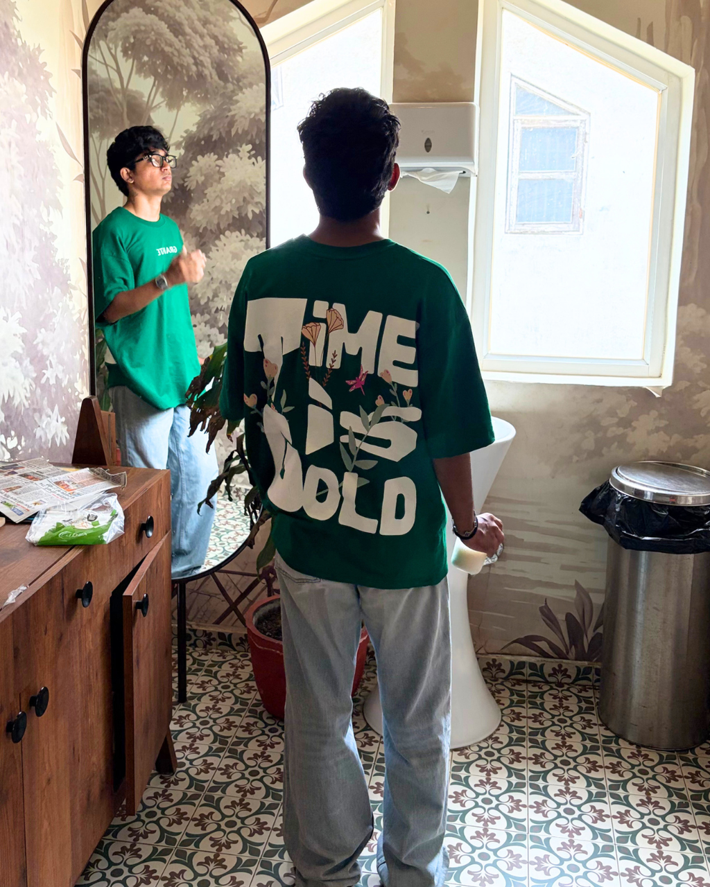A person standing in a bathroom wearing a green oversized T-shirt with a large white graphic on the back that reads 'TIME IS GOLD', paired with light gray sweatpants. The person is shown from behind, with their reflection visible in a large arched mirror on the wall.