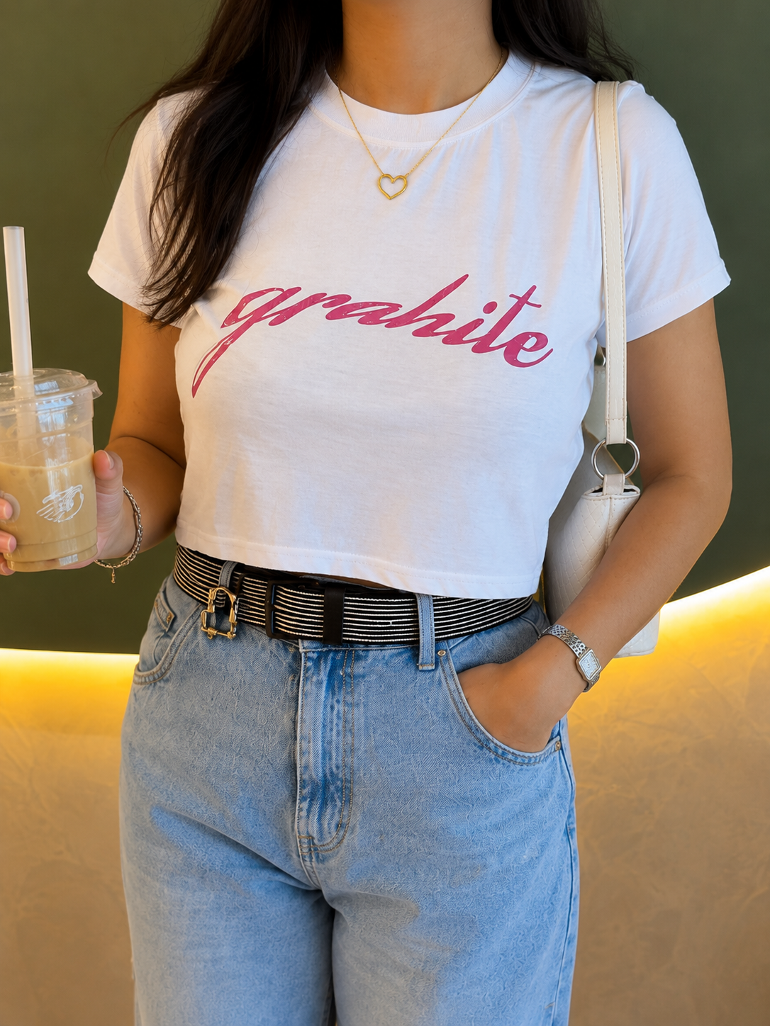 A woman wearing a white cropped t-shirt with 'grahite' printed in pink script across the chest, paired with light blue jeans and a black-and-white striped belt.