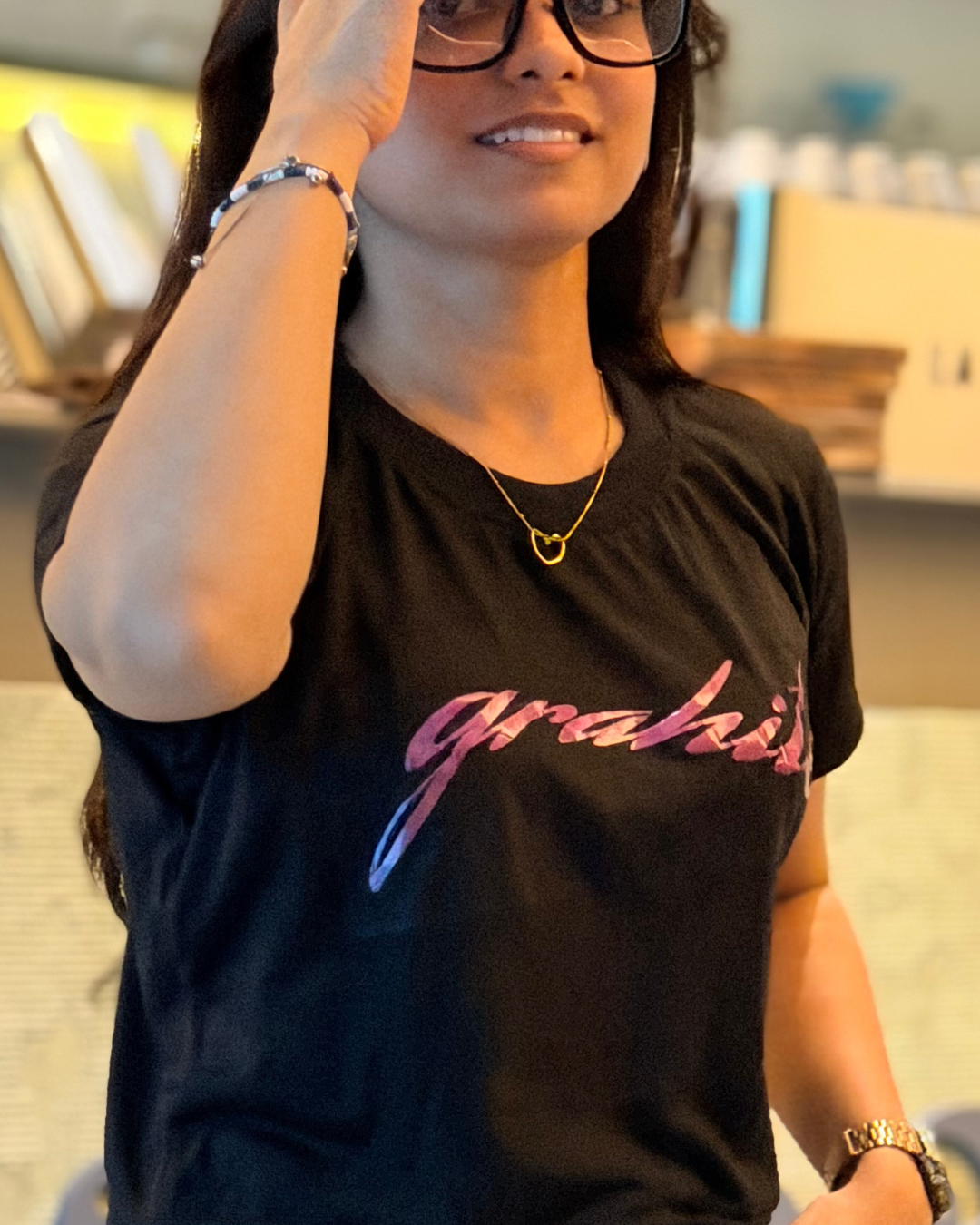 A woman wearing a black CROP TOP with a pink and blue gradient script text that reads 'grahit' across the chest, paired with a gold heart necklace and glasses.