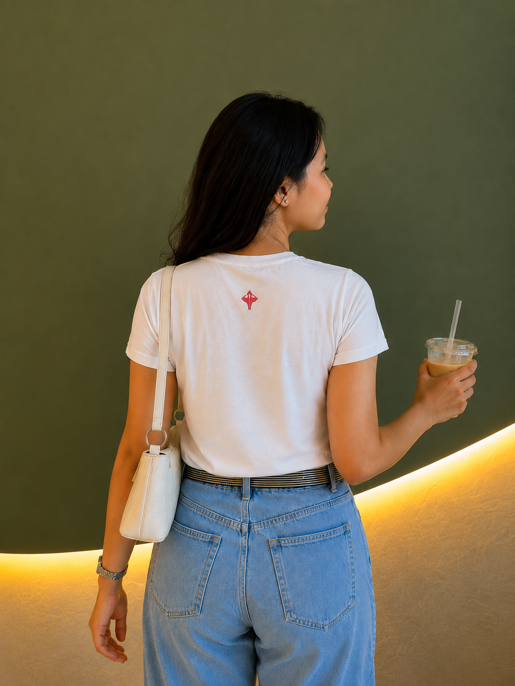A woman viewed from behind wearing a white short-sleeve t-shirt with a small red geometric logo on the upper back, paired with light blue jeans and carrying a white shoulder bag. She is holding an iced coffee against a dark olive green wall with warm yellow lighting.
